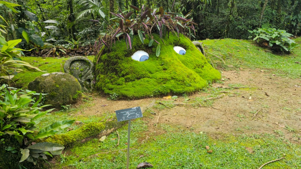 Escultura de musgo verde em formato de rosto com olhos azuis e plantas roxas como cabelo, em meio a uma floresta tropical exuberante com árvores e folhagens densas