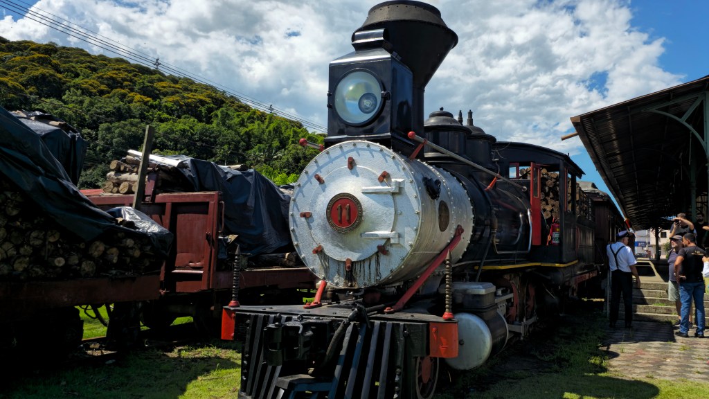 Locomotiva a vapor preta e prateada, número 11 em destaque, estacionada ao lado de vagões com toras de madeira cobertas por lona preta. Ao fundo, uma montanha verdejante e céu azul com nuvens. À direita, pessoas observam a locomotiva sob uma estrutura de estação