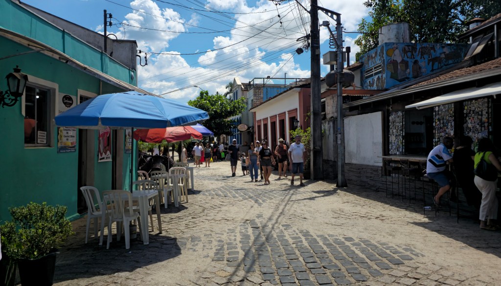 Rua de paralelepípedos com pessoas caminhando sob céu azul com nuvens. À esquerda, um prédio verde-água com mesas e cadeiras brancas sob guarda-sóis azuis e vermelhos. À direita, um bar com balcão e pessoas sentadas, e um prédio azul com grafite de figuras. Postes com fios elétricos e vegetação complementam a cena.