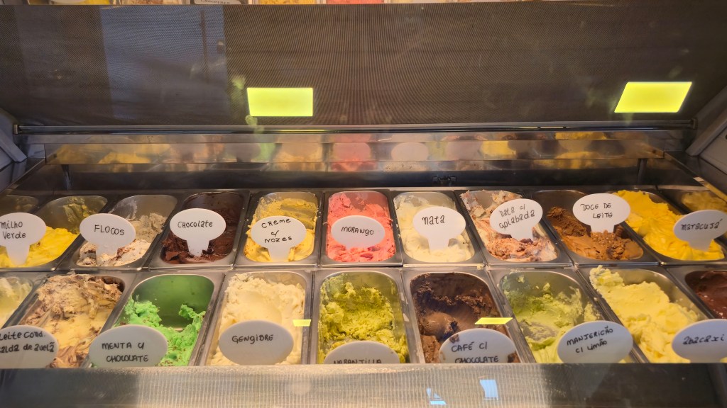 Expositor de sorvetes com 18 sabores em bandejas metálicas, incluindo milho verde, flocos, chocolate, creme com nozes, morango, nata, nata com goiabada, doce de leite, maracujá, leite condensado com calda de queijo, menta com chocolate, gengibre, amarantilha, café com chocolate, manjericão com limão e abacaxi