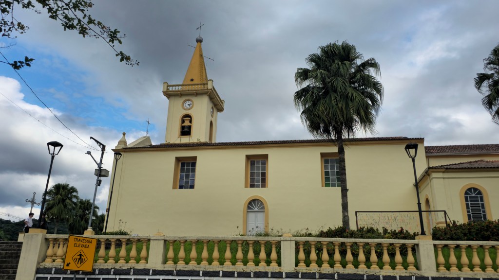 Igreja de cor creme com torre amarela e relógio, sob céu nublado. À direita, uma palmeira alta. Em primeiro plano, um muro com balaustrada amarela e uma placa de trânsito amarela indicando TRAVESSIA ELEVADA. Postes de luz e vegetação complementam a cena.