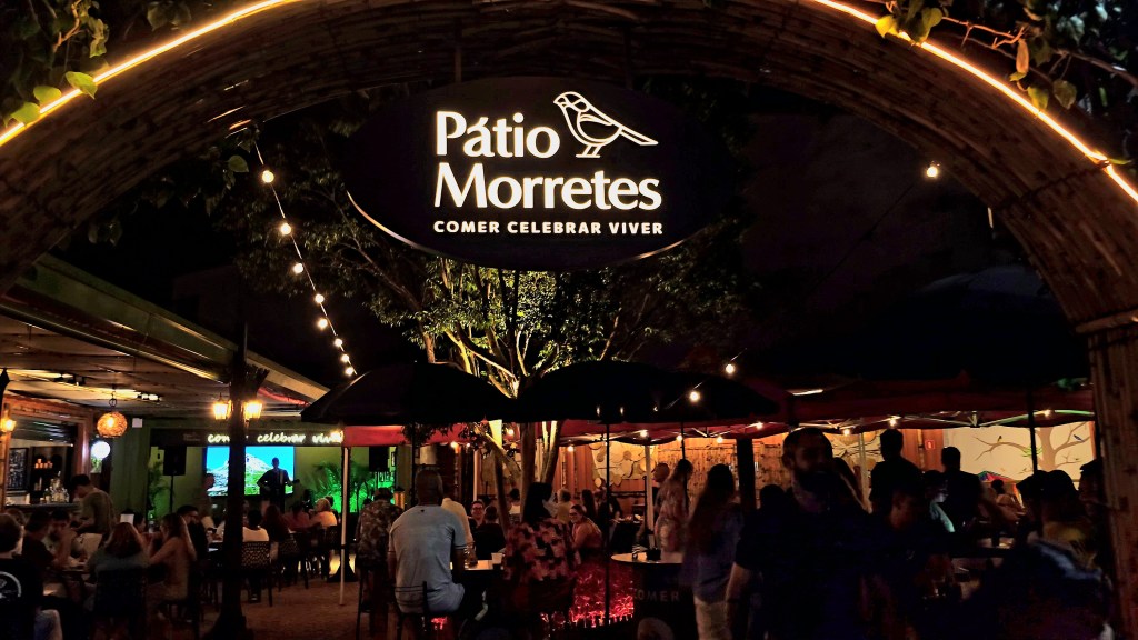 Entrada de um bar noturno com arco de madeira iluminado por luzes amarelas e folhagens. No centro, um letreiro oval com Pátio Morretes e um pássaro, seguido de COMER CELEBRAR VIVER. Pessoas sentadas em mesas e balcões, algumas sob guarda-sóis, com um palco ao fundo exibindo um músico e uma paisagem