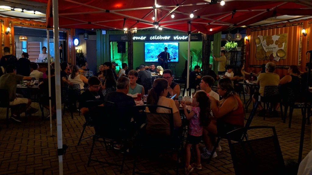 Pessoas sentadas em mesas de um restaurante ao ar livre sob uma tenda vermelha iluminada, com um músico tocando violão no palco ao fundo, onde se lê comer celebrar viver