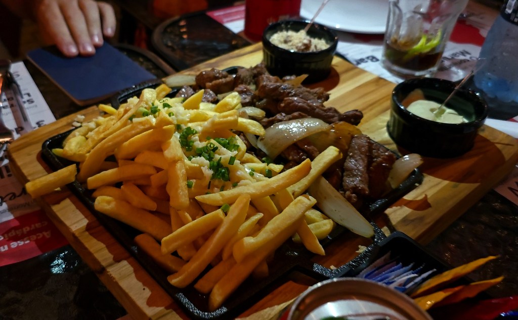 Porção de batatas fritas com queijo e cebolinha, e carne acebolada em chapa preta sobre tábua de madeira, com molhos e farofa em potes pretos, em mesa de restaurante