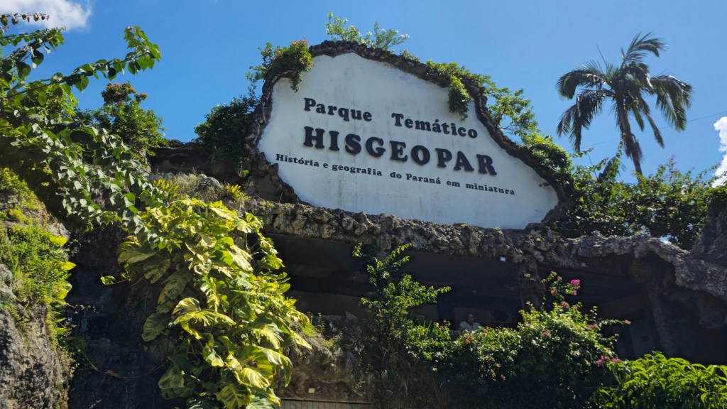 Placa de entrada do Parque Temático HISGEOPAR, com a frase História e geografia do Paraná em miniatura, em uma estrutura rochosa coberta por vegetação verde e exuberante, sob um céu azul com nuvens brancas. Uma palmeira alta se destaca à direita