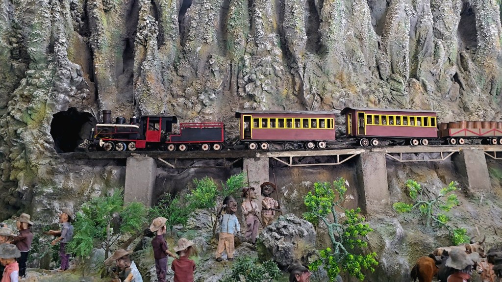 Maquete de trem a vapor preto e vermelho saindo de um túnel na montanha, passando por uma ponte com vagões de passageiros marrons e amarelos. Abaixo, bonecos de pessoas e animais em um cenário rochoso com vegetação
