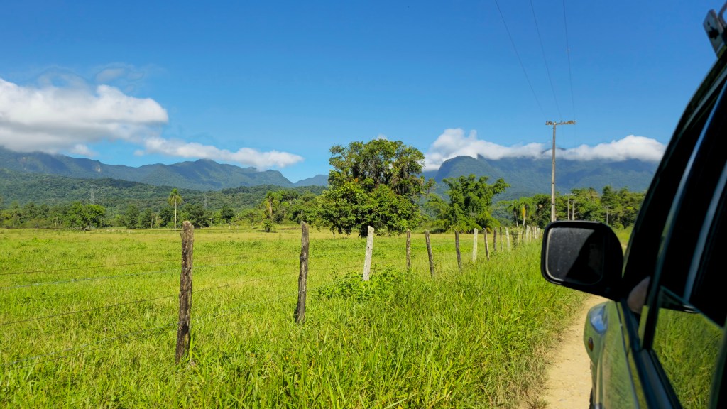 Vista lateral de um carro em uma estrada de terra, com um campo verde e uma cerca de arame à esquerda. Ao fundo, montanhas cobertas por vegetação e um céu azul com nuvens esparsas. Um poste de energia e árvores completam a paisagem rural