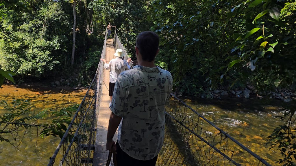 Três pessoas atravessam uma ponte pênsil de madeira e cordas sobre um rio de águas claras e rasas, cercado por mata densa sob sol forte