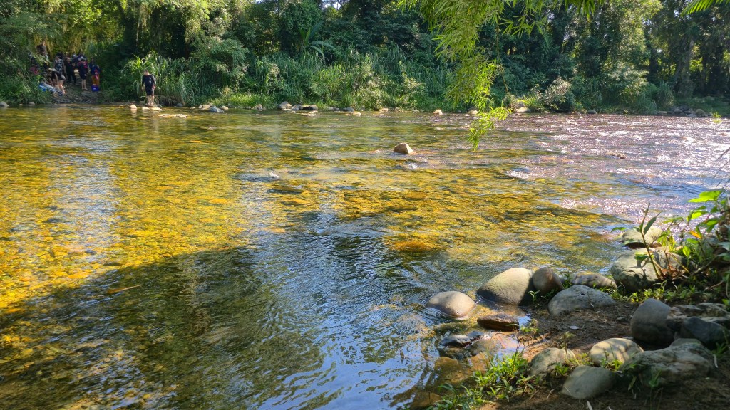 Rio de águas claras e rasas com pedras visíveis no fundo, refletindo o sol. À esquerda, um grupo de pessoas na margem, e um homem de preto em pé sobre as pedras. A margem é densamente arborizada com vegetação verde exuberante
