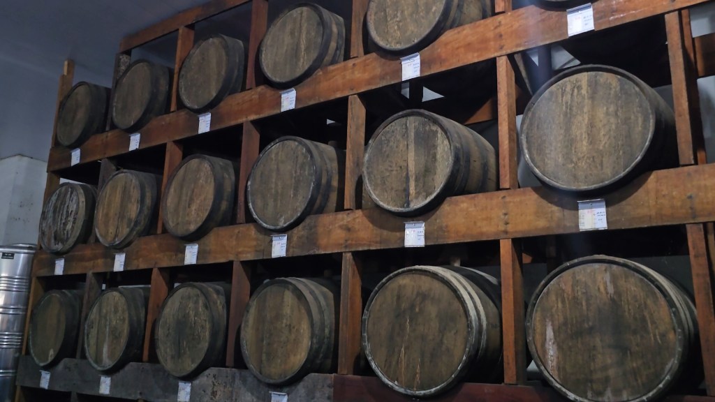 Várias barricas de madeira escura, com aros metálicos, empilhadas em prateleiras de madeira rústica, preenchendo um ambiente de armazenamento. Algumas barricas têm etiquetas brancas presas à madeira. No canto inferior esquerdo, há um empilhamento de recipientes metálicos prateados