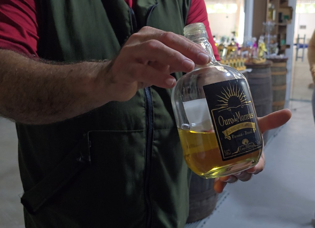 Homem de camisa vermelha e colete verde escuro segura uma garrafa de cachaça Ouro de Morretes com líquido dourado, em ambiente de adega com barris ao fundo
