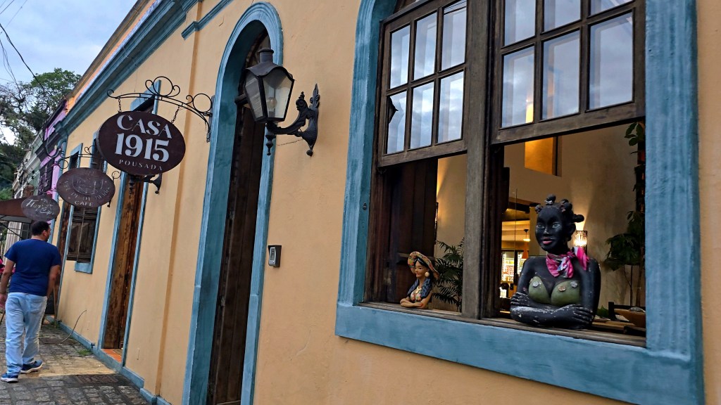 Fachada de uma pousada com paredes amarelas e detalhes azuis. Um homem de camiseta azul e jeans caminha na calçada. Na janela, estátuas de mulheres, uma delas com pele escura, turbante e lenço rosa. Placas de Casa 1915 Pousada e Hot Café penduradas.
