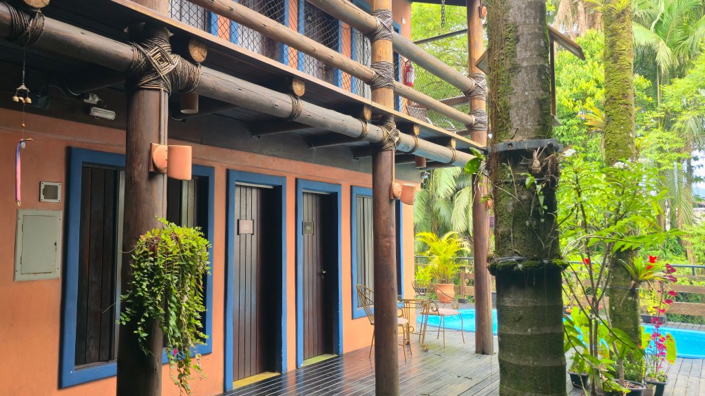 Varanda de pousada rústica com paredes cor de pêssego, portas e janelas com molduras azuis. Há um corrimão de madeira e plantas penduradas. À direita, uma piscina azul e vegetação tropical exuberante.