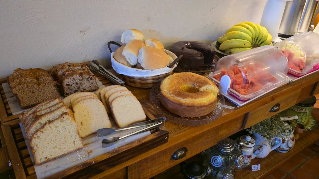 Mesa de café da manhã com pães fatiados, pães franceses em cesto, bolo de chocolate, bolo de fubá, bananas e melancia picada em potes