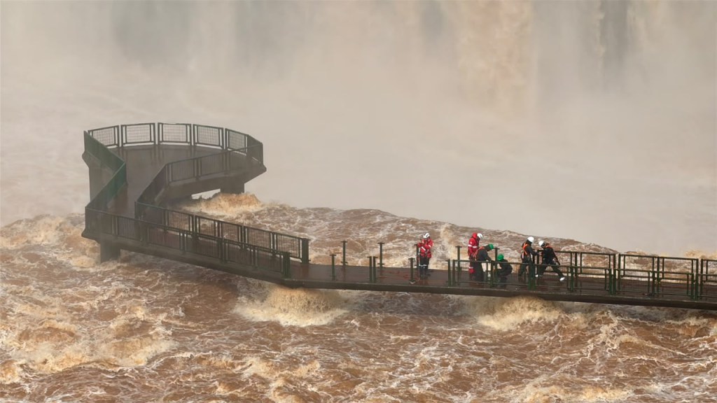 Passarela de metal cinza com corrimão verde sobre rio de águas marrons agitadas, com névoa ao fundo. Seis pessoas, algumas de capacete e colete salva-vidas, estão na passarela, que leva a uma plataforma de observação circular.