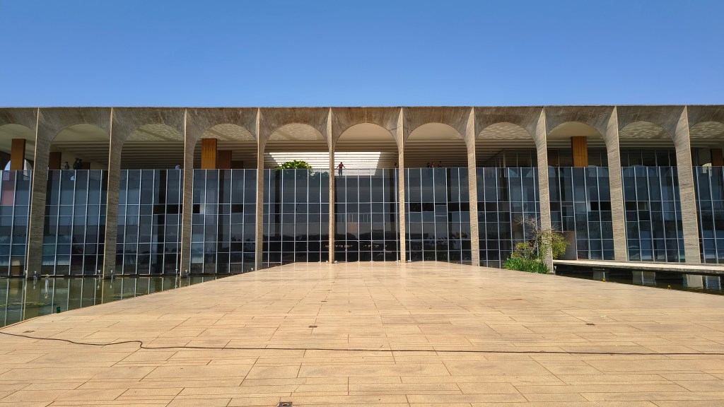 Fachada do Palácio do Itamaraty em Brasília, com arcos de concreto aparente sobre uma parede de vidro espelhado. Um espelho dágua reflete a arquitetura, e um piso de lajotas claras se estende em primeiro plano sob um céu azul claro. Uma pessoa de camisa vermelha observa da varanda superior.