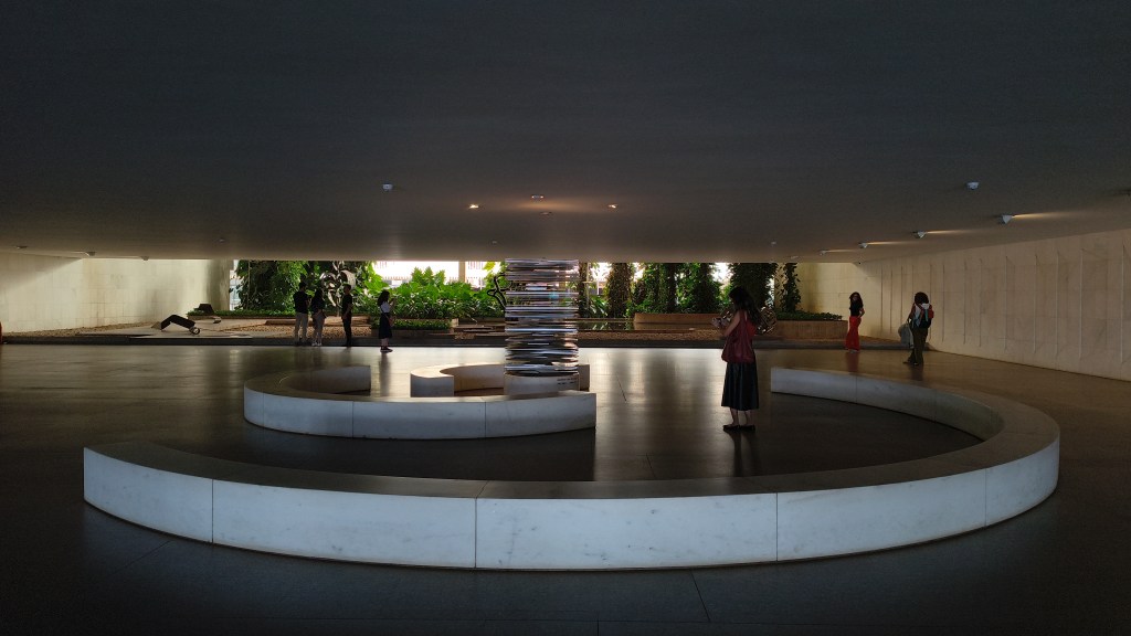Pessoas observam uma escultura cilíndrica de metal espelhado no centro de um piso escuro, cercada por bancos curvos de mármore branco. Ao fundo, um jardim exuberante com árvores e folhagens verdes é visível através de uma abertura retangular, iluminado pela luz natural. O teto baixo e escuro contrasta com a iluminação do jardim e dos bancos.