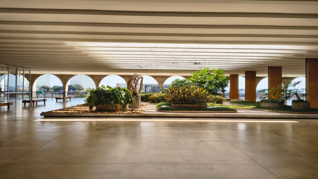 Um jardim interno com plantas verdes e arbustos coloridos, em um ambiente com teto de concreto e vigas horizontais. Ao fundo, arcos de concreto revelam o exterior com prédios e céu azul. O chão é de mármore claro e reflete a luz solar, criando um ambiente sereno e arquitetônico