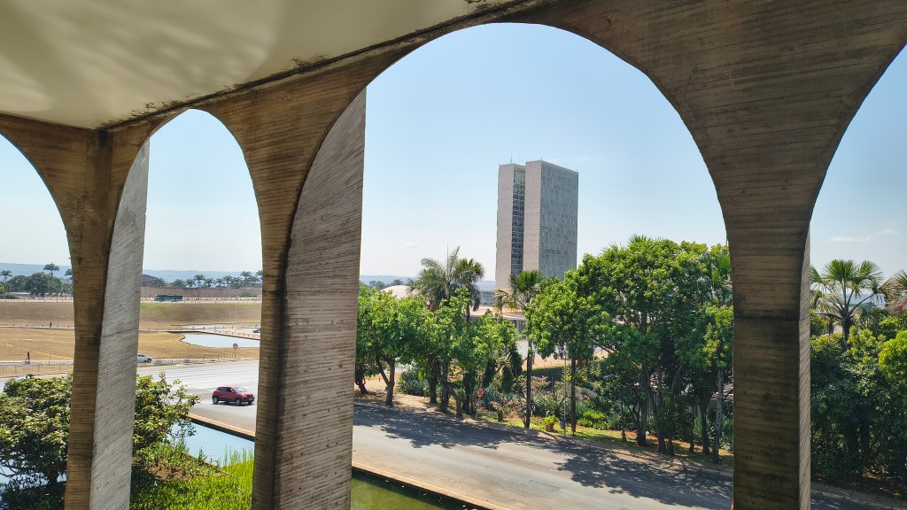 Vista de Brasília através de arcos de concreto, mostrando o anexo do Congresso Nacional, uma rua com um carro vermelho, vegetação exuberante e um céu azul claro
