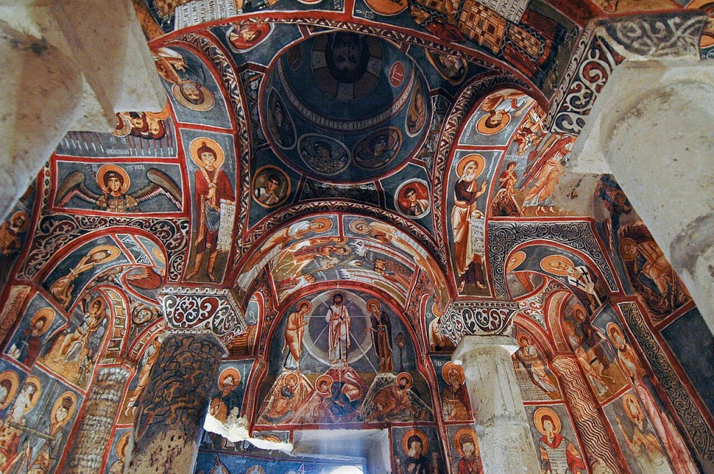 Interior de uma igreja antiga com afrescos bizantinos coloridos no teto e paredes, retratando figuras religiosas em tons de vermelho, azul, dourado e branco, com detalhes ornamentados. Uma cúpula central e arcos exibem cenas bíblicas e santos, enquanto pilares de pedra rústica sustentam a estrutura, revelando a arte desgastada pelo tempo
