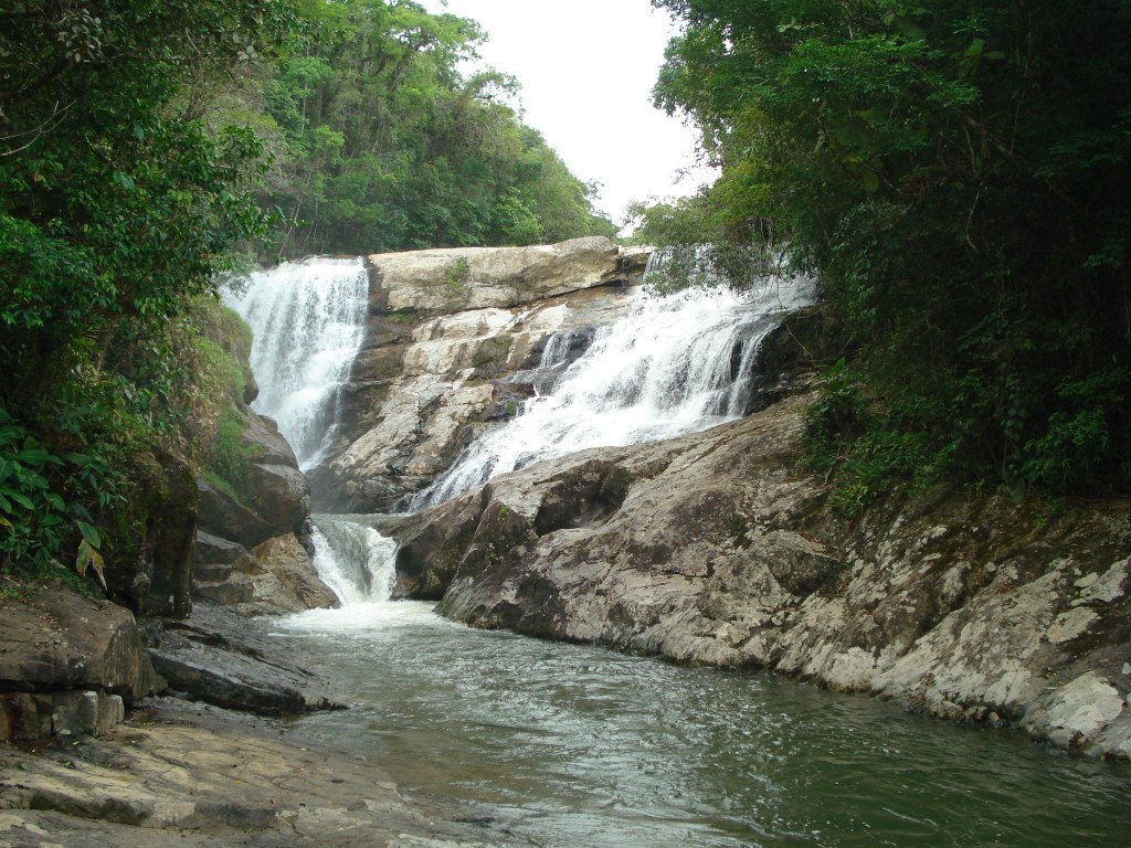 Cachoeira com quedas dágua em rochas escuras e claras, cercada por vegetação densa e verde, com um rio fluindo em primeiro plano