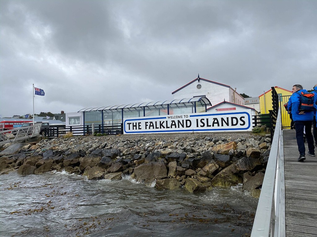 Pessoas caminhando em um píer de madeira, com um letreiro branco e azul Welcome to The Falkland Islands em primeiro plano. Ao fundo, construções coloridas e a bandeira das Ilhas Falkland tremulando sob um céu nublado, com o mar agitado por pedras na margem
