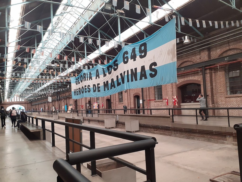 Bandeira argentina com a frase Gloria a los 649 héroes de Malvinas pendurada em um galpão de tijolos com teto de vidro e bandeirinhas, pessoas caminham e sentam em bancos