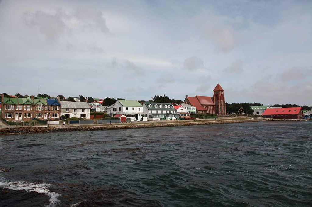 Vista costeira de uma cidade com casas coloridas e uma igreja de telhado vermelho, sob céu nublado. O mar agitado, com algas escuras, ocupa o primeiro plano.