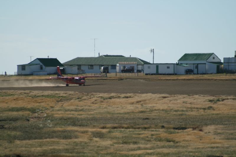 Pequeno avião vermelho e branco decolando em pista de terra, levantando poeira, com casas brancas de telhado verde ao fundo em um dia ensolarado