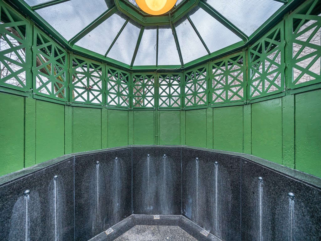 Interior de um gazebo octogonal verde com teto de vidro e uma luminária amarela central. A parte inferior é de granito escuro com detalhes metálicos e manchas claras de água escorrendo