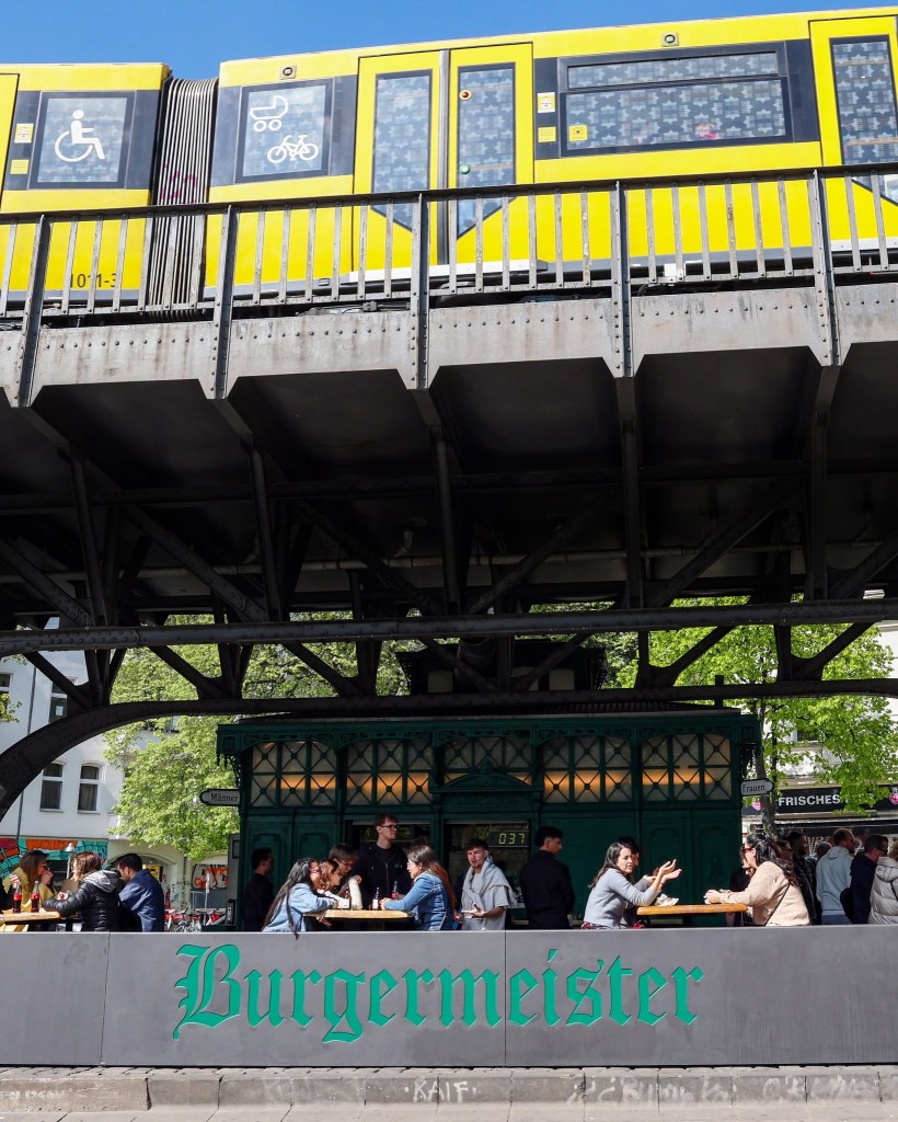 Um trem amarelo passa sobre uma estrutura elevada de metal, enquanto pessoas sentam em mesas de madeira sob a estrutura, em frente a um quiosque verde. A palavra Burgermeister está em destaque na parte inferior da imagem