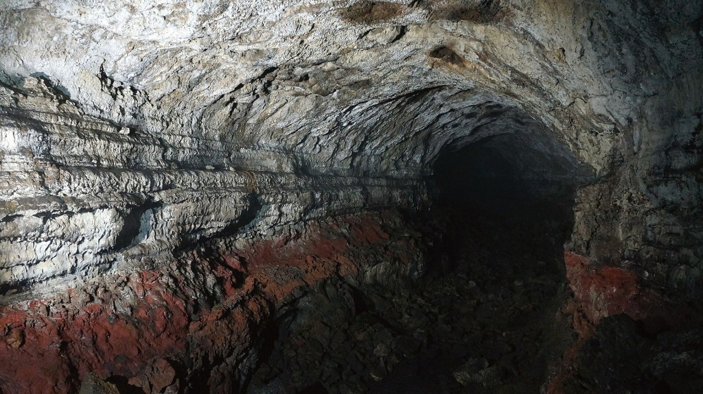 Interior de uma caverna escura com paredes rochosas irregulares em tons de cinza e branco, refletindo a luz. Uma faixa de rocha avermelhada se estende na parte inferior da parede esquerda, e o chão é escuro e rochoso. O túnel se aprofunda na escuridão ao fundo