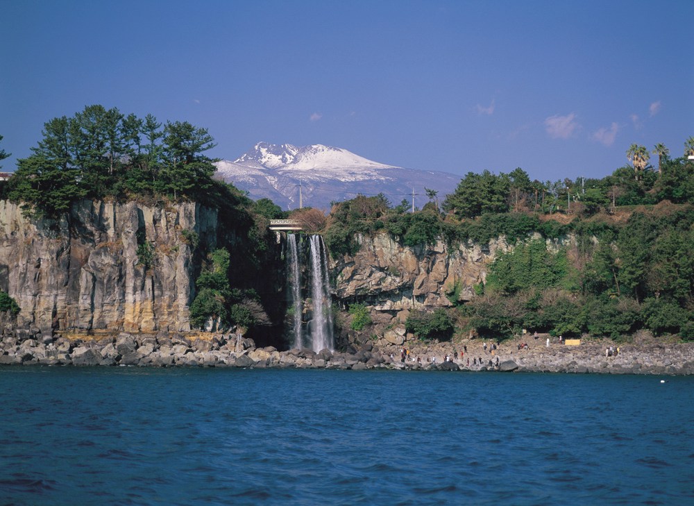 Cachoeira em Jeju, Coreia do Sul, com água caindo de um penhasco rochoso para o mar azul. Pessoas observam na margem rochosa, e ao fundo, uma montanha com pico nevado sob céu claro