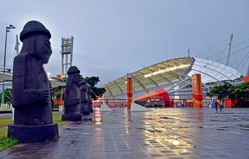 Estátuas de pedra escuras, chamadas Dol Hareubang, alinhadas em uma praça molhada, com um estádio moderno de teto branco e laranja ao fundo, sob um céu nublado