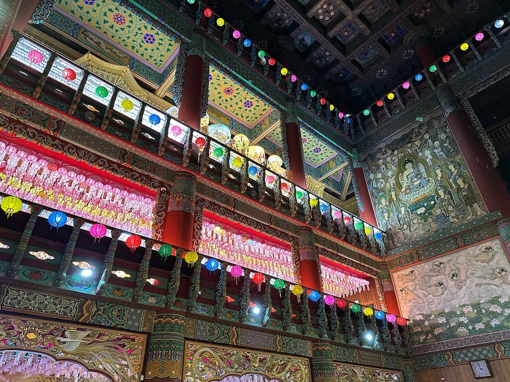 Interior de um templo budista ricamente decorado com lanternas coloridas penduradas em fileiras. As paredes e o teto exibem pinturas detalhadas e padrões geométricos em tons vibrantes de verde, vermelho, azul e dourado. Uma grande pintura mural de figuras divinas adorna a parede direita, enquanto o teto tem vigas ornamentadas.