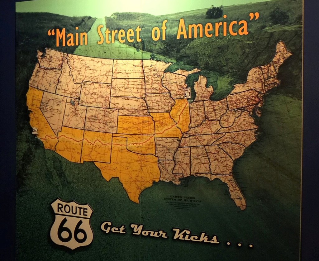 Mapa dos Estados Unidos com a Rota 66 destacada em amarelo, atravessando o país. O título Main Street of America aparece no topo, e o logotipo Route 66 Get Your Kicks está na parte inferior esquerda. O fundo é verde escuro, simulando uma paisagem montanhosa