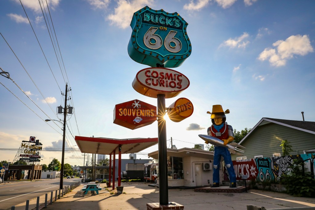 Placa vintage azul e branca da Rota 66 com letreiros neon Bucks on 66, Cosmic Curios, Souvenirs e Toys em um poste. Ao lado, uma estátua de astronauta-caubói segurando um foguete, em frente a um prédio com grafite. O sol brilha atrás dos letreiros, criando um reflexo. Ao fundo, uma rua com postes de luz e um céu azul com nuvens esparsas