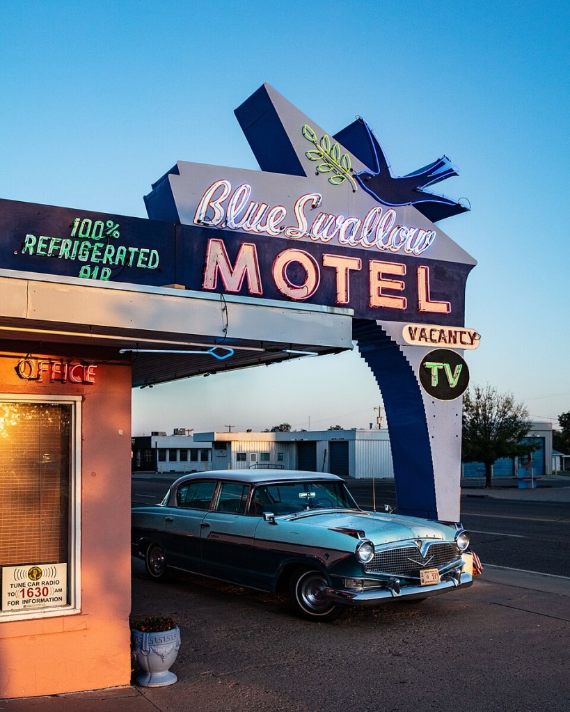 Letreiro neon azul e rosa do Blue Swallow Motel com uma andorinha azul e um carro antigo azul-claro estacionado em frente ao escritório cor de pêssego, sob um céu azul claro