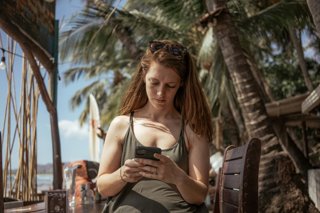 Uma mulher confere o celular sentada em um banco sob palmeiras, em um dia ensolarado