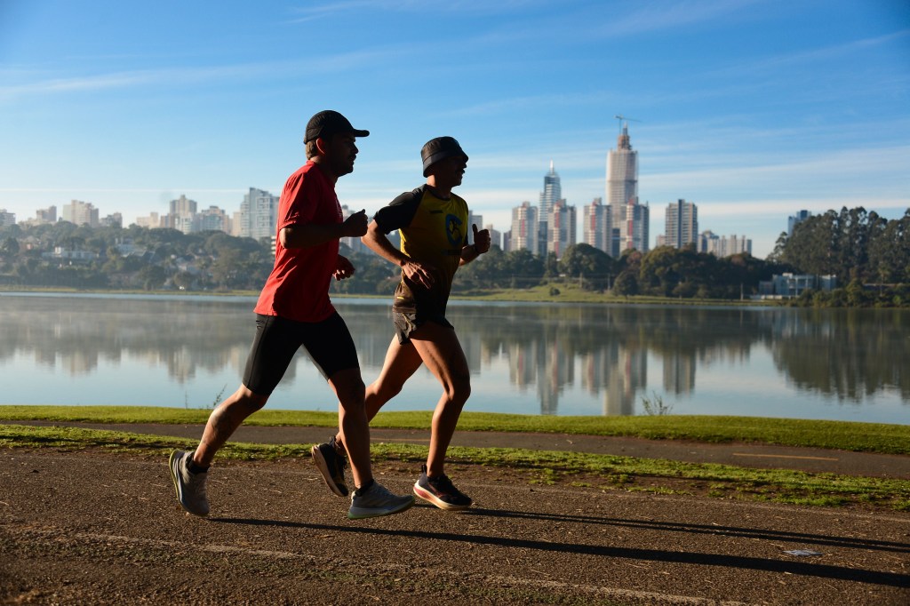 Dois homens correm em uma pista de terra ao lado de um lago, com a silhueta de uma cidade ao fundo. O homem da frente veste camiseta vermelha e boné preto, o de trás usa camiseta amarela e chapéu. O céu está azul e o sol ilumina a cena, refletindo os prédios na água.