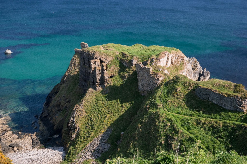 Ruínas de um castelo antigo no topo de um penhasco rochoso coberto por grama verde, com o mar azul-turquesa e azul-escuro ao fundo e uma praia de pedras na base