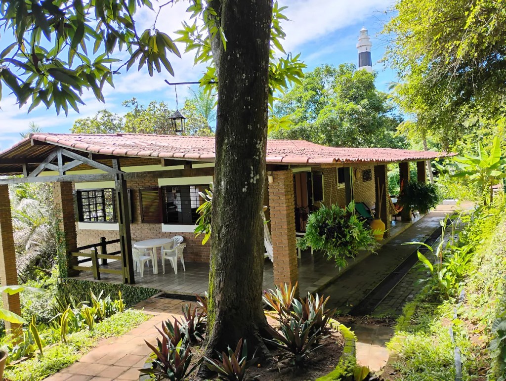 Casa de tijolos com telhado de cerâmica e varanda, cercada por vegetação exuberante. Um farol branco e preto se destaca ao fundo, sob céu azul com nuvens. Uma mesa e cadeiras brancas estão na varanda, e um caminho de tijolos leva à entrada. A cena é iluminada pelo sol, transmitindo tranquilidade