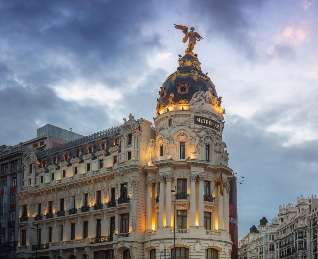 Edifício Metrópolis em Madrid, com fachada branca ornamentada e cúpula escura dourada, coroada por uma estátua alada. As janelas estão iluminadas com luzes amareladas, sob um céu nublado de fim de tarde