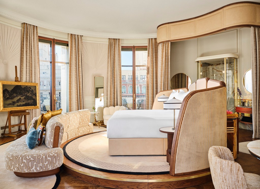 Quarto de hotel luxuoso com cama king-size de dossel bege, sofá curvo e poltrona estofados em tons claros, e uma pintura em cavalete. O ambiente tem janelas altas com cortinas douradas, espelho de corpo inteiro e piso de madeira com tapete redondo central. A decoração é sofisticada, com detalhes em madeira e iluminação suave