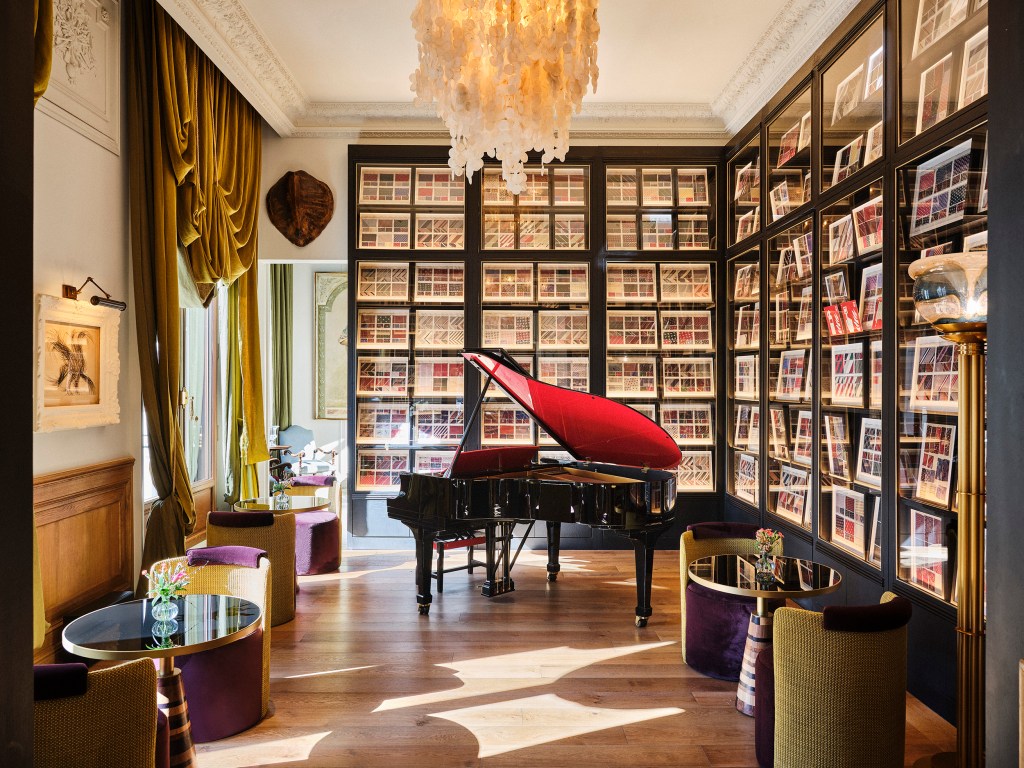 Sala elegante com piano de cauda preto e tampa vermelha, rodeado por estantes escuras com molduras de gravuras. Cortinas douradas e poltronas roxas e amarelas complementam o ambiente, iluminado por um lustre de conchas e luz natural no chão de madeira