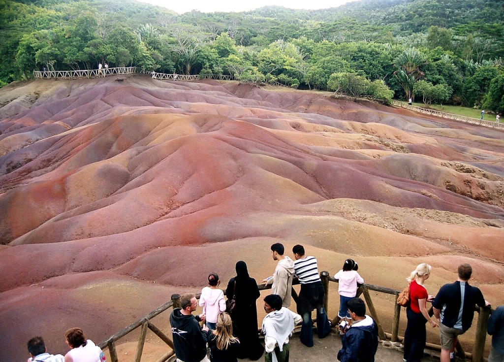 Turistas observam as Dunas de Sete Cores em Chamarel, Ilhas Maurício, com colinas onduladas em tons de marrom, roxo e laranja. Um grupo de pessoas está em primeiro plano, em uma plataforma de madeira, e outras caminham em passarelas elevadas ao fundo, cercadas por vegetação densa e montanhas