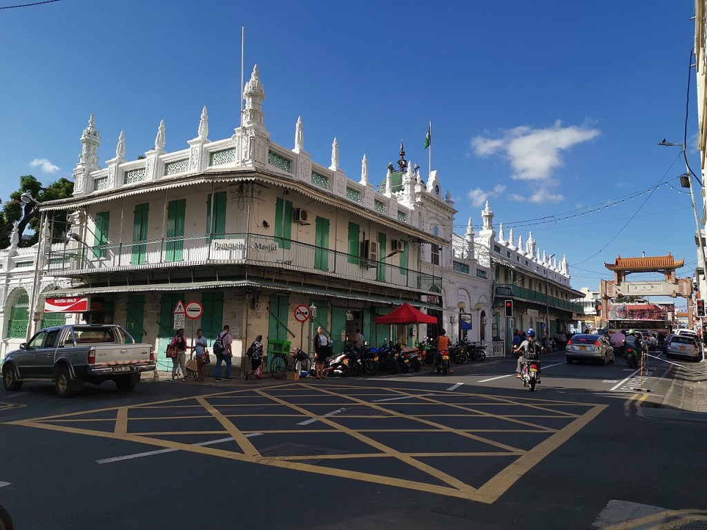 Rua movimentada em Port Louis, Maurício, com a mesquita Jummah Masjid de arquitetura colonial branca e venezianas verdes. Pessoas caminham na calçada, carros e motos circulam, e ao fundo, um portal chinês tradicional marca a entrada de um bairro. O céu está azul com poucas nuvens
