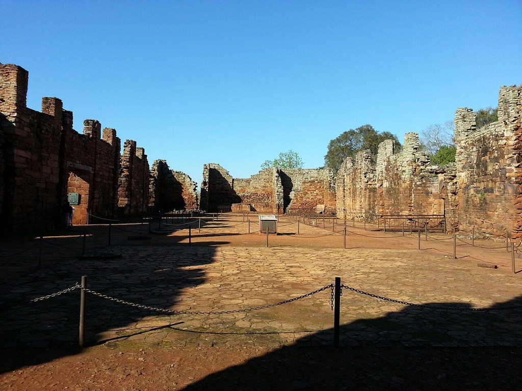 Ruínas de San Ignacio Miní, com paredes de tijolos avermelhados e pedras, sob céu azul claro. O chão é de terra batida e pedras, com correntes baixas delimitando caminhos. Sombras alongadas cobrem parte do solo, e árvores verdes aparecem ao fundo