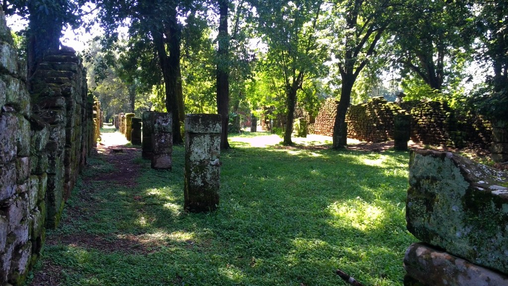 Ruínas históricas com paredes de pedra cobertas por musgo, ladeando um caminho de terra batida. À direita, um gramado verde com árvores altas e frondosas, e mais ruínas ao fundo. A luz do sol filtra-se pelas folhas, criando sombras no chão.