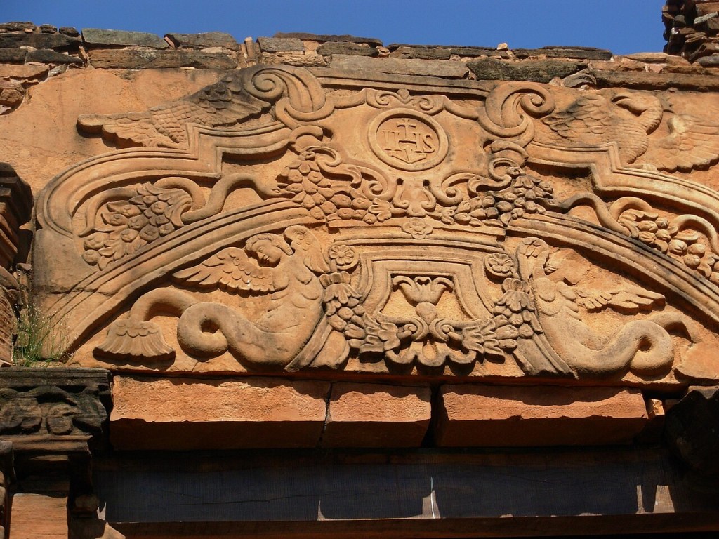 Detalhe de um entalhe em pedra arenito com relevos barrocos. No centro superior, um círculo com as letras IHS. Abaixo, dois anjos alados com caudas de peixe, rodeados por folhagens e flores. A cor da pedra é marrom-avermelhada, com partes desgastadas e musgo. O céu azul claro aparece ao fundo